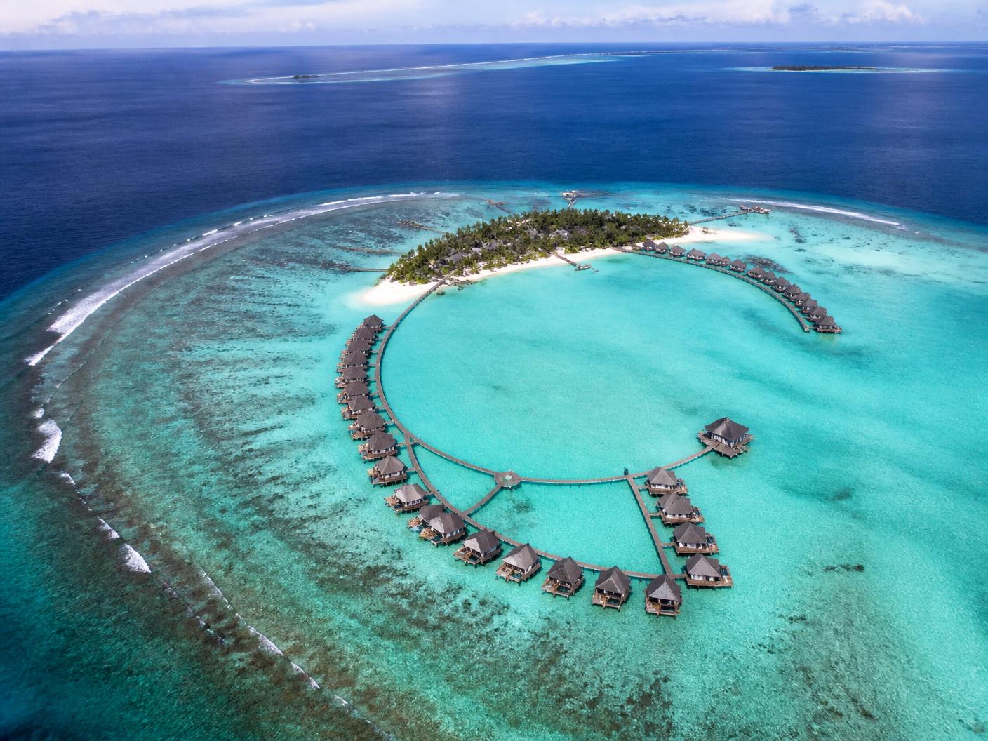 Aerial view of a Maldivian atoll with a curving row of overwater villas above a pale turquoise lagoon