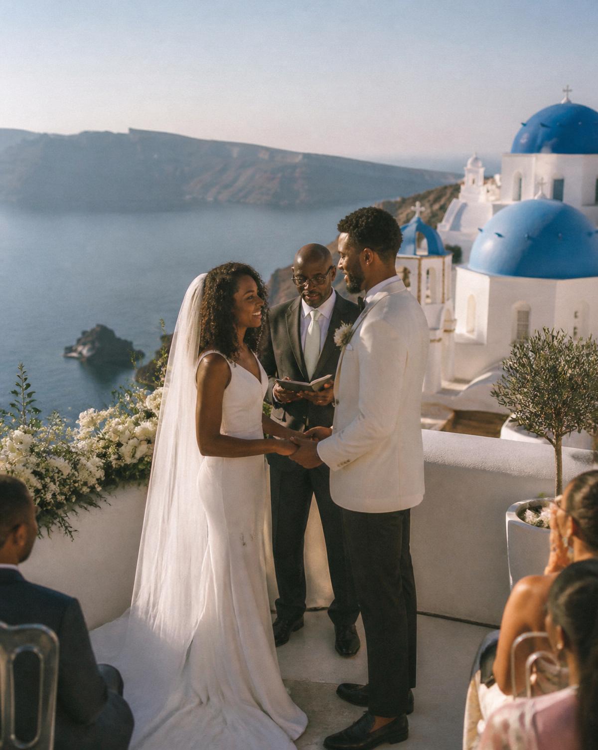 A Black bride and groom exchange vows on a whitewashed clifftop terrace in Santorini overlooking the Aegean caldera